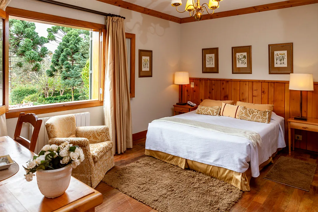 Quarto com paredes de madeira, cama de roupa dourada e vista de araucária